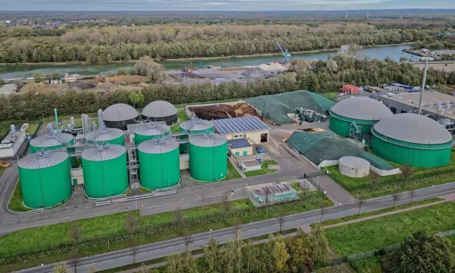 Grüne Biogas-Behälter und große Gasspeicher prägen diese Anlage, die die **Biogas Zukunft** symbolisiert. Im Hintergrund ein Fluss und bewaldete Ufer.