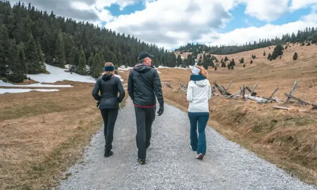 Trzy osoby idące ścieżką w górach. W tle las i pagórkowaty krajobraz.