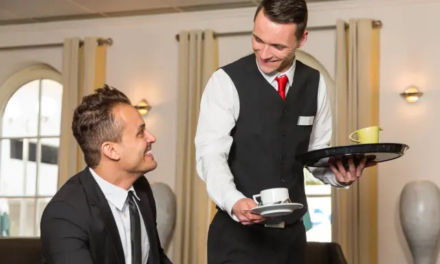 Un serveur souriant présente une tasse de café à un client, illustrant le protocole de service à table.