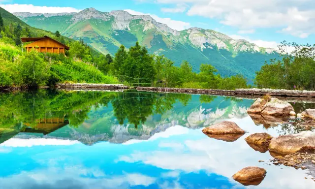 Drewniany domek na zielonym zboczu, odbijający się w spokojnej tafli wody. Idealne miejsce na odpoczynek po górskiej wędrówce, gdzie ubezpieczenie górskie zapewni spokój.