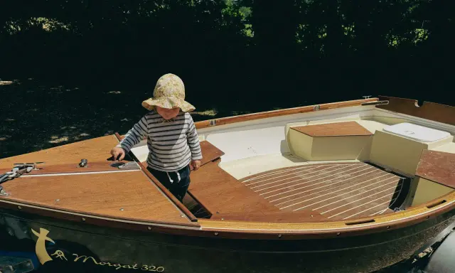 Un enfant en chapeau fleuri explore l'intérieur d'un bateau en bois, le Morgat 320, prêt pour l'aventure.