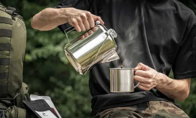 Mężczyzna nalewa kawę do kubka z metalowego czajniczka. Obok leży plecak i paczki kawy. Idealny **aeropress przepis** na kempingu.