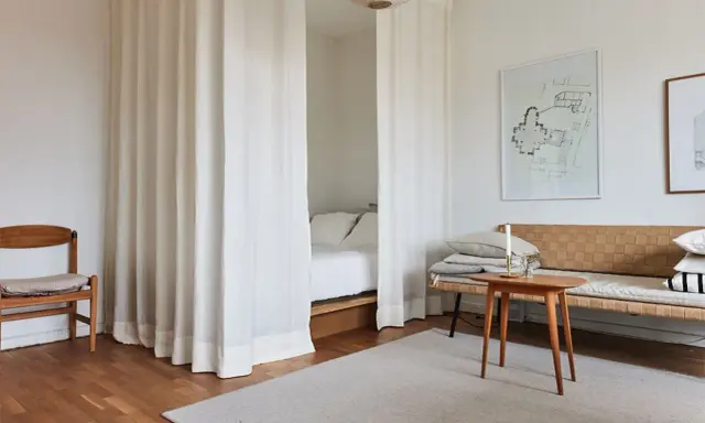 Vita draperier skärmar av ett rum och skapar en mysig sovalkov. En trästol och en soffa med kuddar står i rummet.