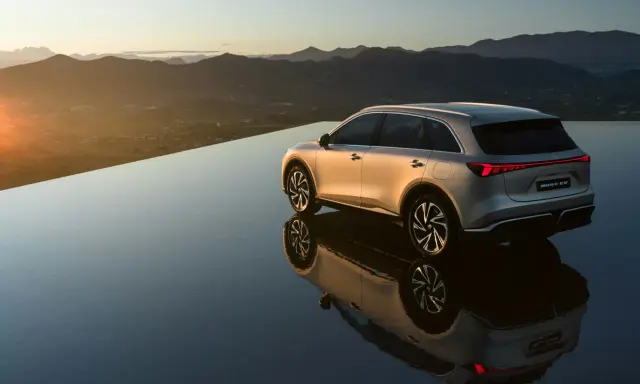 El MG6 EV plateado se refleja en una superficie de agua infinita al atardecer, con montañas al fondo.