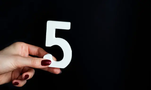 Dłoń z ciemnymi paznokciami trzyma białą cyfrę 5, symbolizującą numerologiczna 5 mężczyzna, na czarnym tle.