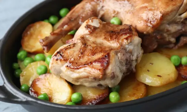 Un saporito coniglio in umido con patate arrosto e piselli verdi, servito in una cocotte scura.