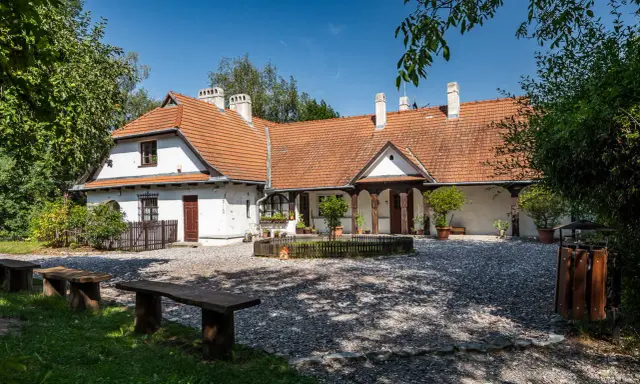 Urokliwy dom z czerwonym dachem w Bronowicach Małych, otoczony zielenią. Widać drewniane ławki i kamienny dziedziniec.