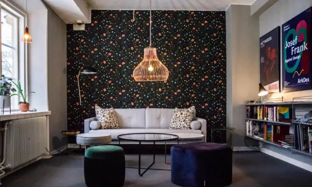En mysig lounge med blommig tapet, en grå soffa, färgglada sittpuffar och en hylla full med böcker. En affisch för "Josef Frank - Against Design" hänger på väggen.