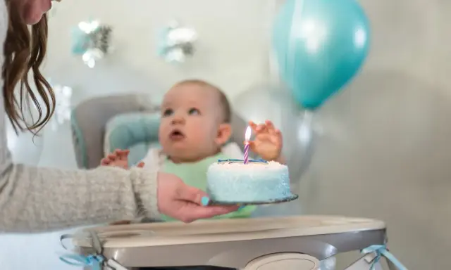 Mama podaje tort z jedną świeczką na roczek maluszkowi. Balony i dekoracje tworzą radosną atmosferę.