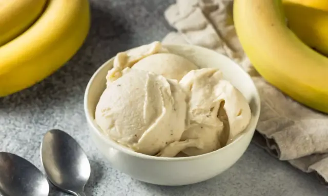 Un cremoso helado de platano servido en un cuenco blanco, con plátanos amarillos frescos al fondo.