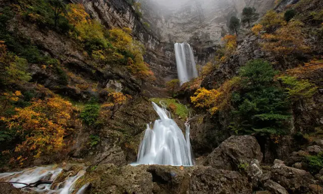 Cascada en un cañón rocoso, con árboles otoñales y niebla. El agua cae como un zafiro lagunazo, creando un espectáculo natural.