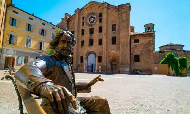 Statua bronzea in una piazza di una città dell'Emilia Romagna, con un'antica chiesa in mattoni sullo sfondo.