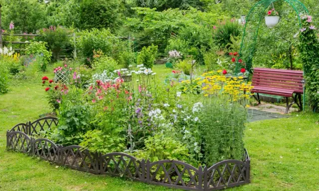 Un exuberante macizo de flores de verano, con rosas rojas, margaritas blancas y flores amarillas vibrantes, bordeado por una valla decorativa.