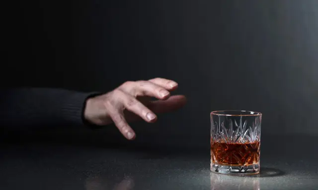 Mano tesa verso un bicchiere di whisky, un'immagine che evoca la domanda: quanto beve un alcolista al giorno?