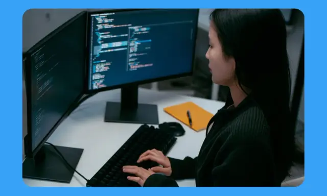 Jeune femme travaillant sur un ordinateur, avec deux écrans affichant du code. Sa position devant l'écran du bureau est concentrée.