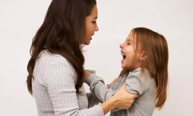 Mama śmieje się, łapiąc córkę za ręce. Dziecko radośnie szarpie się w nerwach, śmiejąc się głośno.