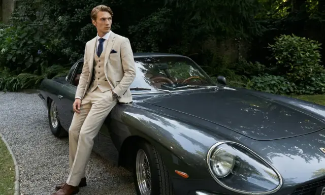 A man in a light-colored suit, perfect for mens summer cocktail attire, leans against a vintage car.