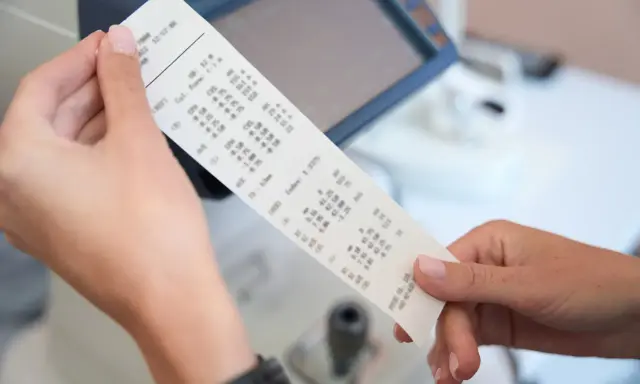 Dłonie trzymają wydruk z danymi optometrycznymi, gdzie widoczny jest numer paragonu.