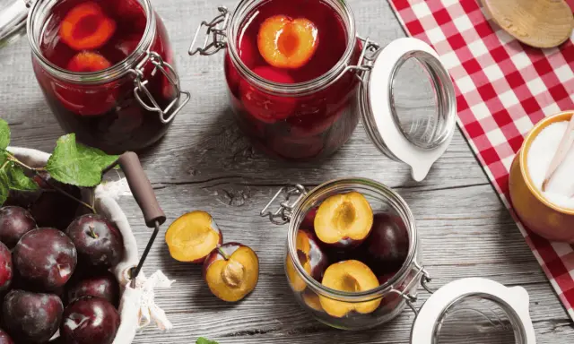 Frische Pflaumen werden für das Einkochen vorbereitet. Ganze und halbierte Früchte liegen neben Gläsern mit eingelegten Pflaumen.