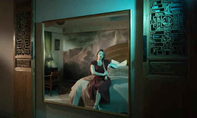Elisa Miller sits on a bed, her reflection captured in a large mirror. The room features ornate wooden panels and a dramatic mountain mural.