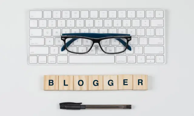 Okulary, klawiatura i drewniane klocki z napisem "BLOGGER" - idealne narzędzia do pisania bloga po angielsku.
