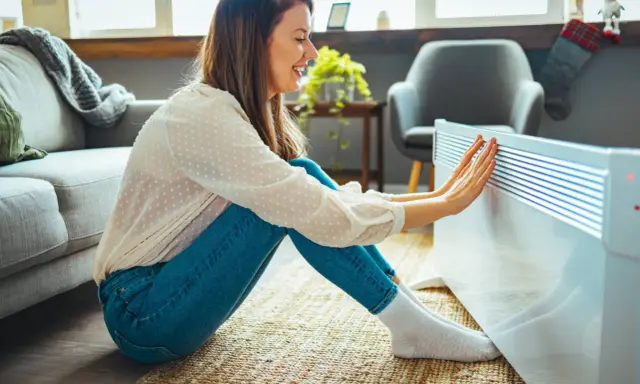 Mujer sonríe mientras calienta sus manos en un radiador, disfrutando de la temperatura ideal en casa en invierno.