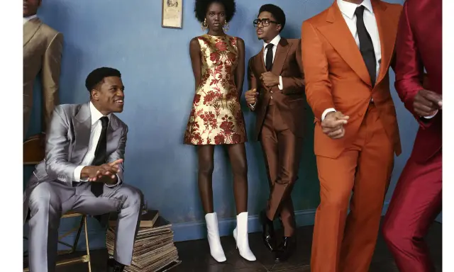 A group of stylish individuals in smart cocktail attire, featuring bold suits and a patterned dress, pose against a blue wall.