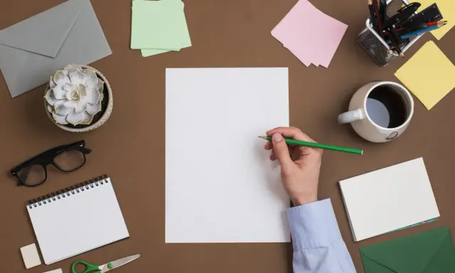 Student pisze list motywacyjny. Na biurku leżą koperty, notatnik, okulary, kubek kawy i przybory do pisania.