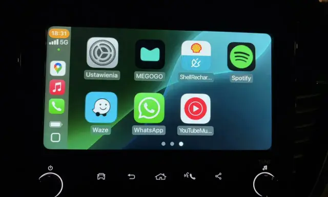 Ekran samochodu z ikonami aplikacji: Ustawienia, MEGOGO, Shell Recharge, Spotify, Waze, WhatsApp, YouTube Music. Działa system iOS CarPlay.