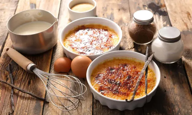Dwa kremowe desery creme brulee z karmelizowanym cukrem, obok składniki: śmietanka, jajka, laski wanilii, cukier.