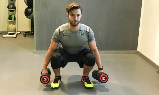 Mann macht Kniebeugen mit Kurzhanteln. Er hält zwei Hanteln mit je 14 kg Gewicht.