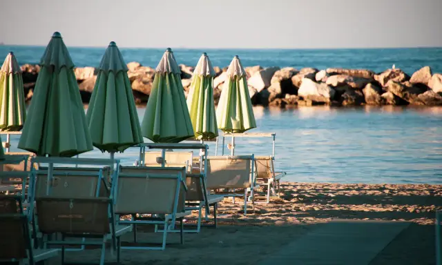 Ombrelloni verdi e lettini vuoti sulla spiaggia di una città riviera romagnola, con il mare calmo e una diga di sassi sullo sfondo.