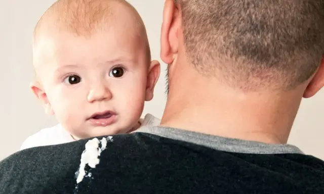 En söt bebis med stora ögon tittar fram över pappans axel. En liten vit fläck syns på pappans tröja, kanske från en rapning.