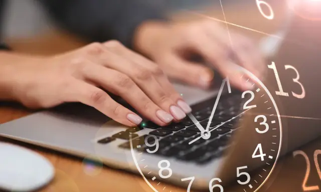 Dłonie piszą na klawiaturze laptopa, a nad nimi unosi się zegar. 0440 znaczenie czasu w pracy.