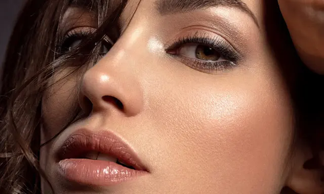 Primo piano di un volto femminile con un sofisticato trucco occhi marroni, labbra luminose e pelle radiosa.