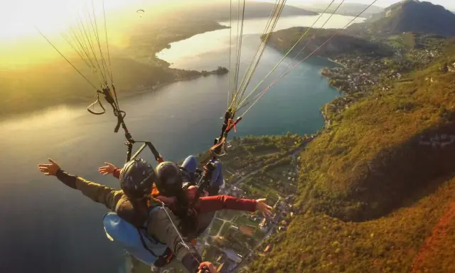 Vol en parapente biplace au-dessus d'un lac, avec un paysage de montagne et de village. Philippe Jeantot, malgré un accident passé, continue de voler.