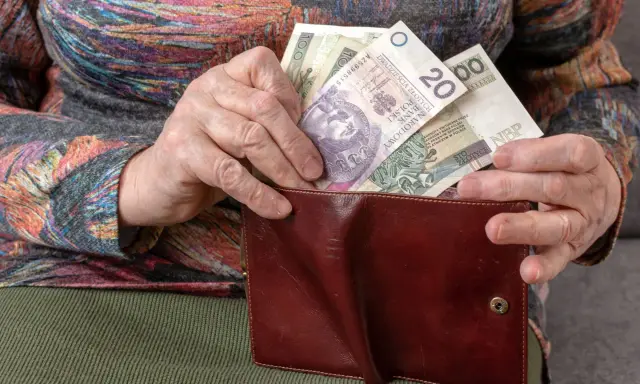 Dłoń starszej osoby wkłada polskie banknoty do brązowego portfela. Widać nominały 20, 100 i 50 złotych, stanowiące ekwiwalent codziennych wydatków.
