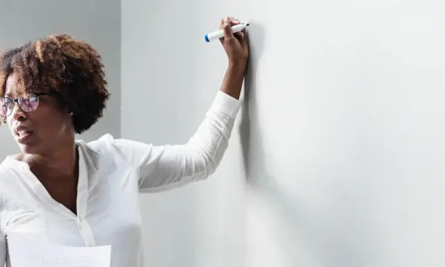 Frau mit Brille und Lockenwicklern hält einen Stift und bietet Nachhilfe an, während sie auf eine weiße Wand blickt.
