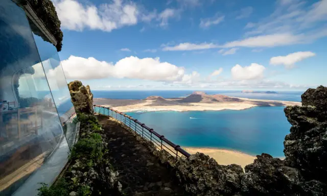 Blick vom Mirador del Río auf das Tal der tausend Palmen Lanzarote. Blaue See, Inseln und eine Hütte mit Glasfront.