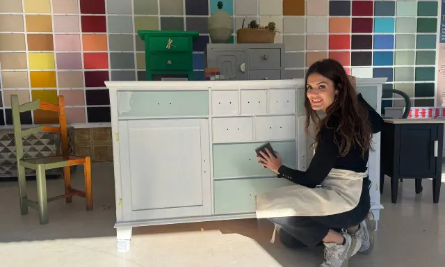 Mujer sonríe mientras trabaja en un mueble, aplicando pintura blanca. Aprende como pintar un mueble oscuro en blanco sin lijar.