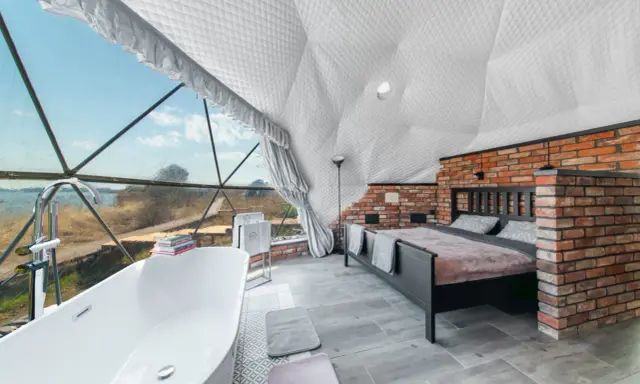 Luksusowy glamping nad jeziorem: wanna z widokiem, łóżko i ceglana ściana w kopułowym wnętrzu.