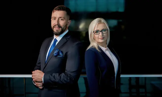 Para prawników w eleganckich garniturach, mężczyzna i kobieta, stojący na tle ciemnego biura.