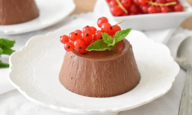 Panna cotta czekoladowa z czerwonymi porzeczkami i miętą na białym talerzyku.