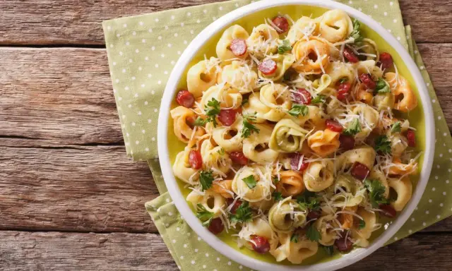 Kolorowa sałatka z tortellini, kawałkami kiełbasy i parmezanem, udekorowana natką pietruszki, podana na talerzu na zielonej serwetce w kropki.