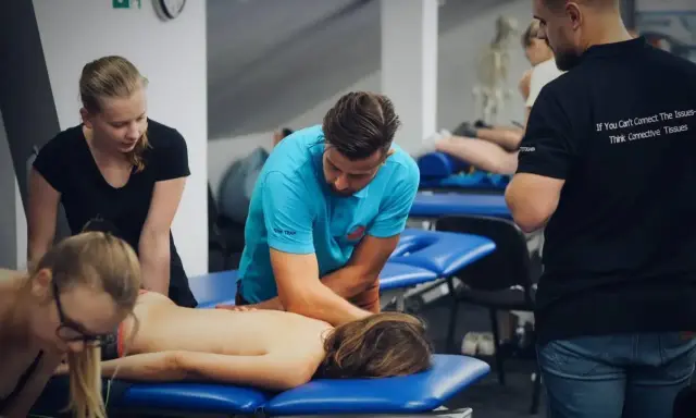 Uczestnicy szkolenia wykonują masaż tkanek miękkich na pacjencie leżącym na kozetce. Terapeuta w niebieskiej koszulce demonstruje technikę.
