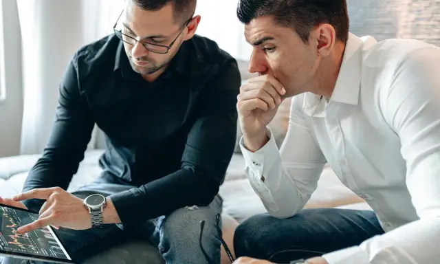 Dos hombres analizan gráficos en una tablet. Uno señala la pantalla, discutiendo cómo domiciliar la nómina para optimizar finanzas.