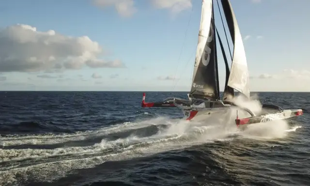 Le trimaran "Actual" d'Yves Le Blevec fend les vagues, propulsé par le vent sous un ciel bleu.