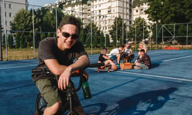 Uśmiechnięty chłopak na rowerze z butelką piwa, obok grupa znajomych. Może to być scena, która zakończy się mandatem za jazdę rowerem po alkoholu.