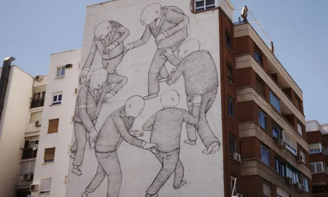A striking street art style mural depicts a group of bald figures in suits, seemingly engaged in a peculiar, interconnected dance or struggle on a building's facade.