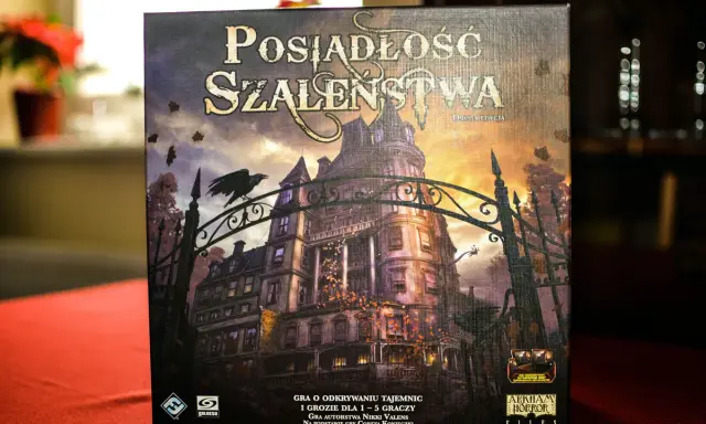 Pudełko gry "Posiadłość Szaleństwa" z mroczną posiadłością i wroną na bramie. Dodatki do tej gry planszowej zapowiadają nowe, przerażające przygody.